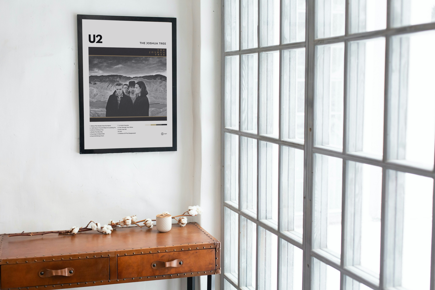 Framed U2 poster on a wall above a wooden desk with a large window in the background.