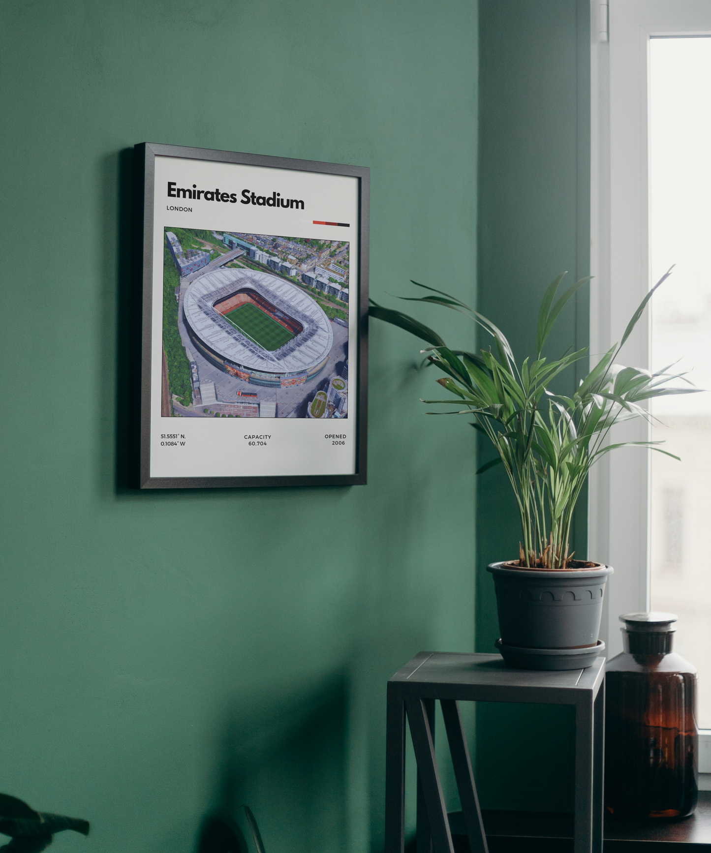 Framed poster of Emirates Stadium on a green wall with a plant and bottle in the foreground.