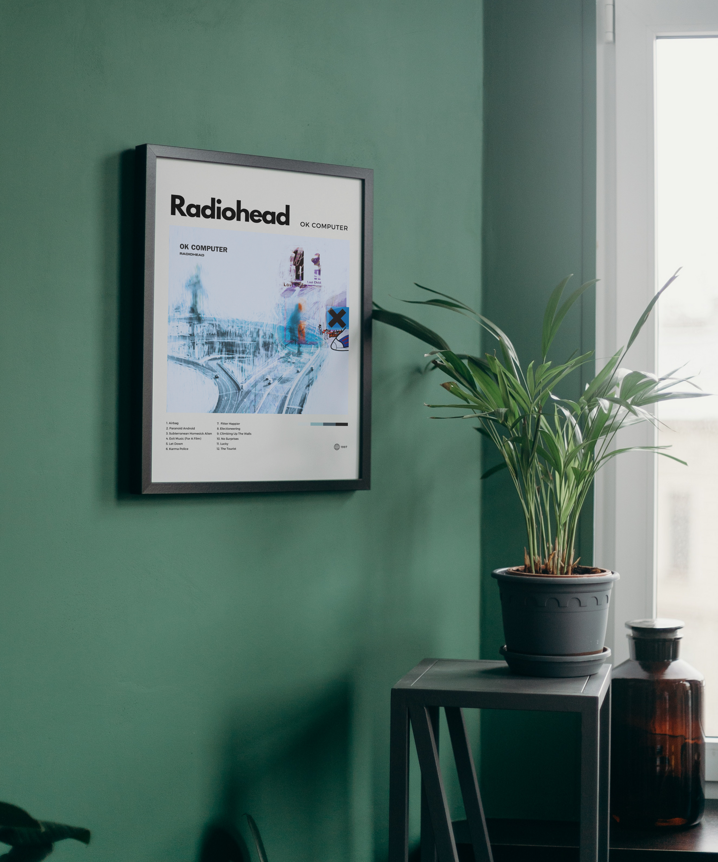 Framed Radiohead album cover on a green wall with a plant and bottle in the foreground