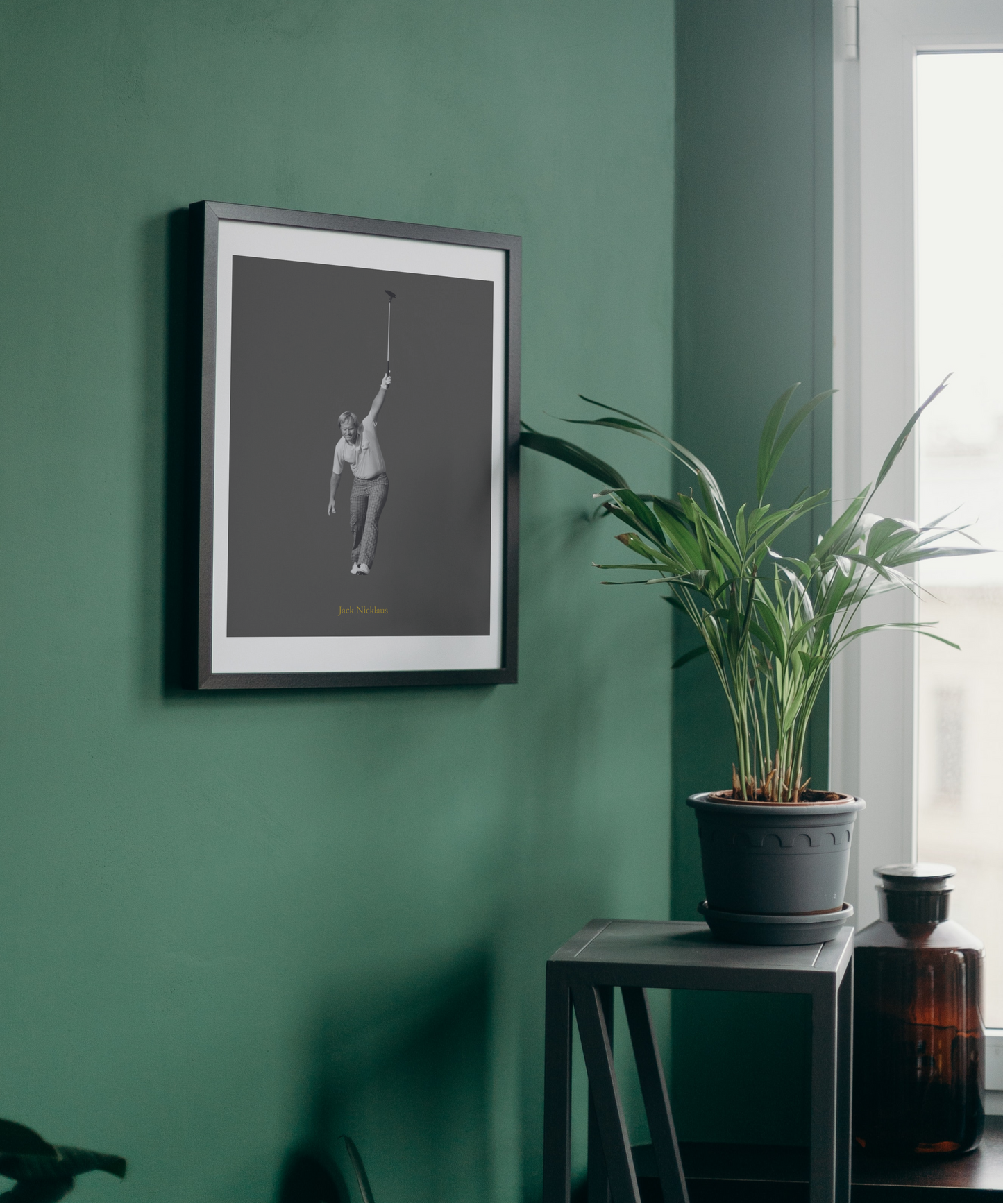 Framed artwork of a Jack Nicklaus playing golf on a green wall with a plant and bottle in the foreground.