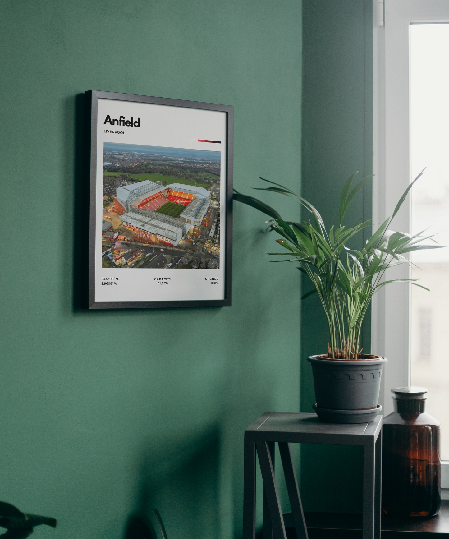 Framed poster of Anfield stadium on a green wall with a plant and bottle in the foreground.