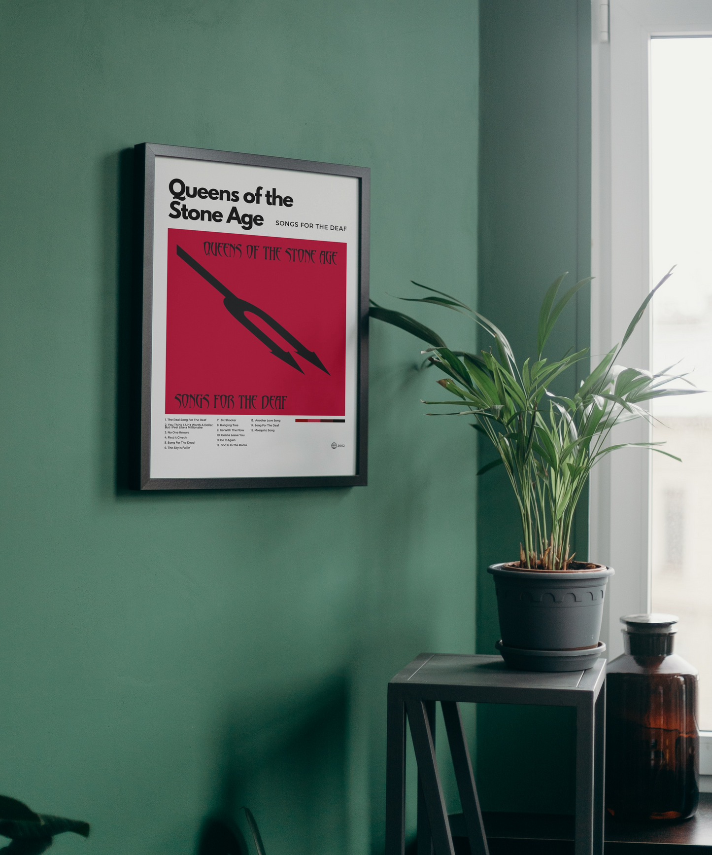 Framed Queens of the Stone Age poster on a green wall with a plant and bottle in the foreground.