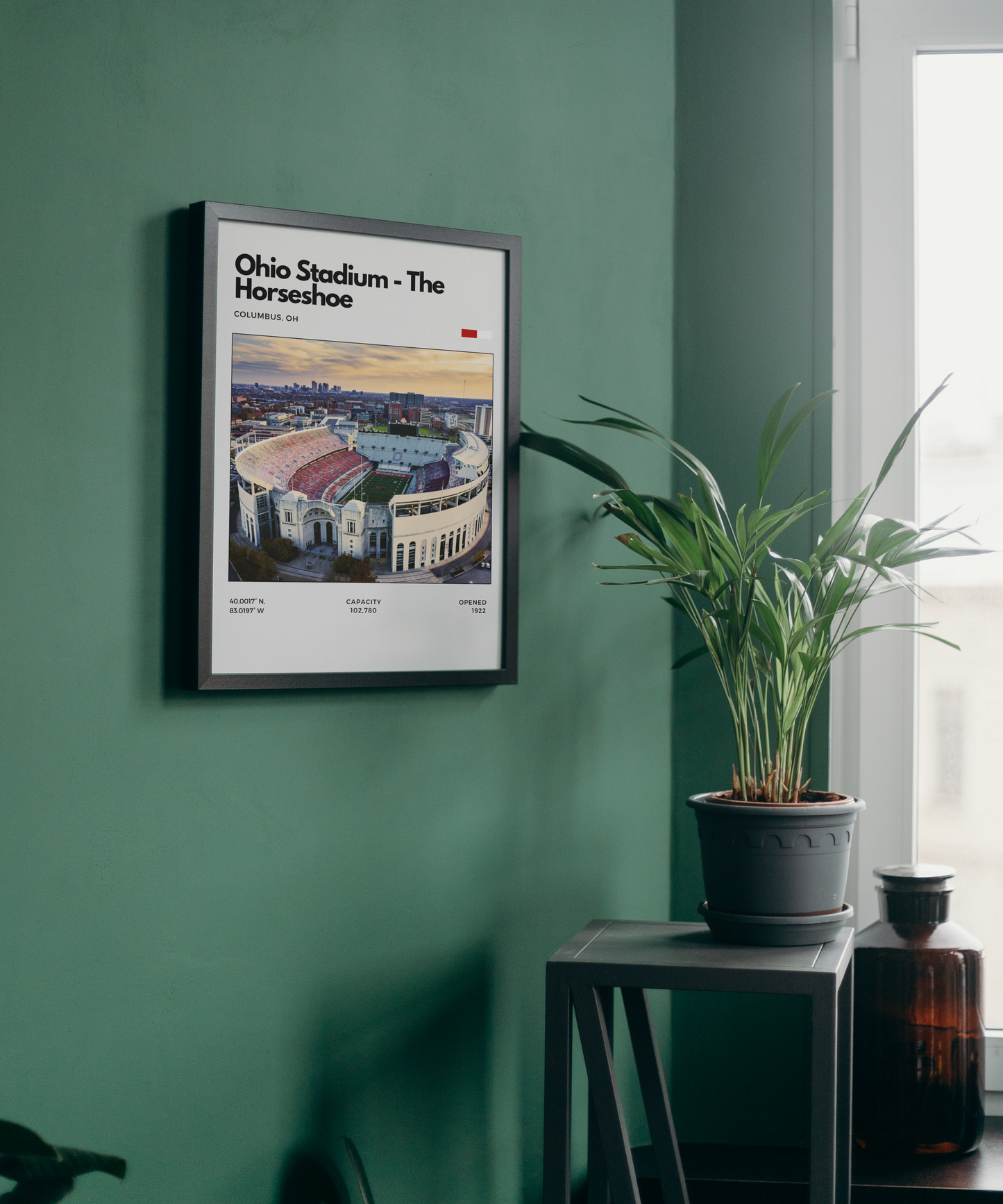 Framed poster of Ohio Stadium on a green wall with a plant and bottle in the foreground.