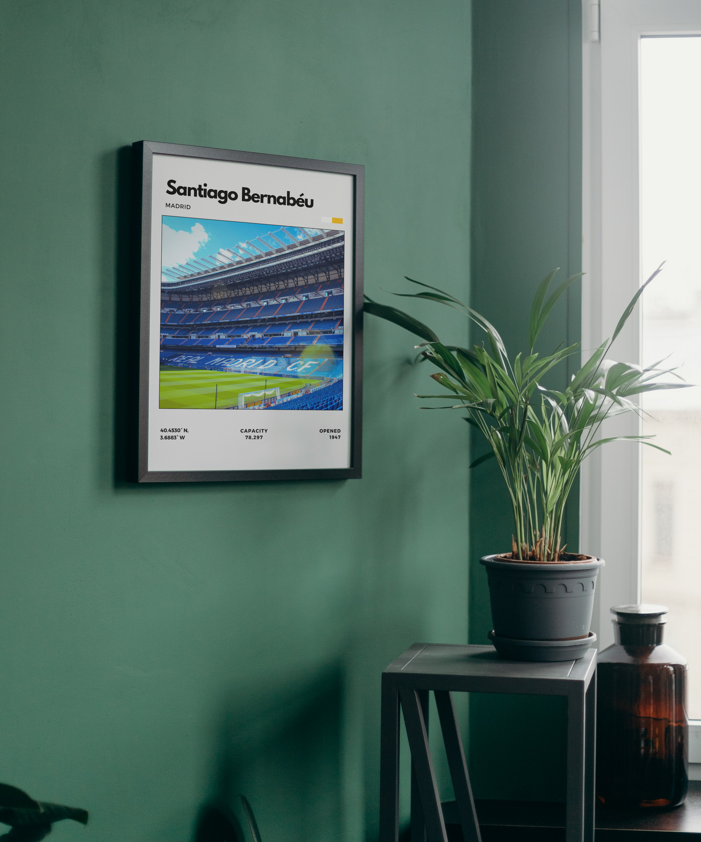 Framed poster of Santiago Bernabéu stadium on a green wall with a plant and bottle in the foreground.