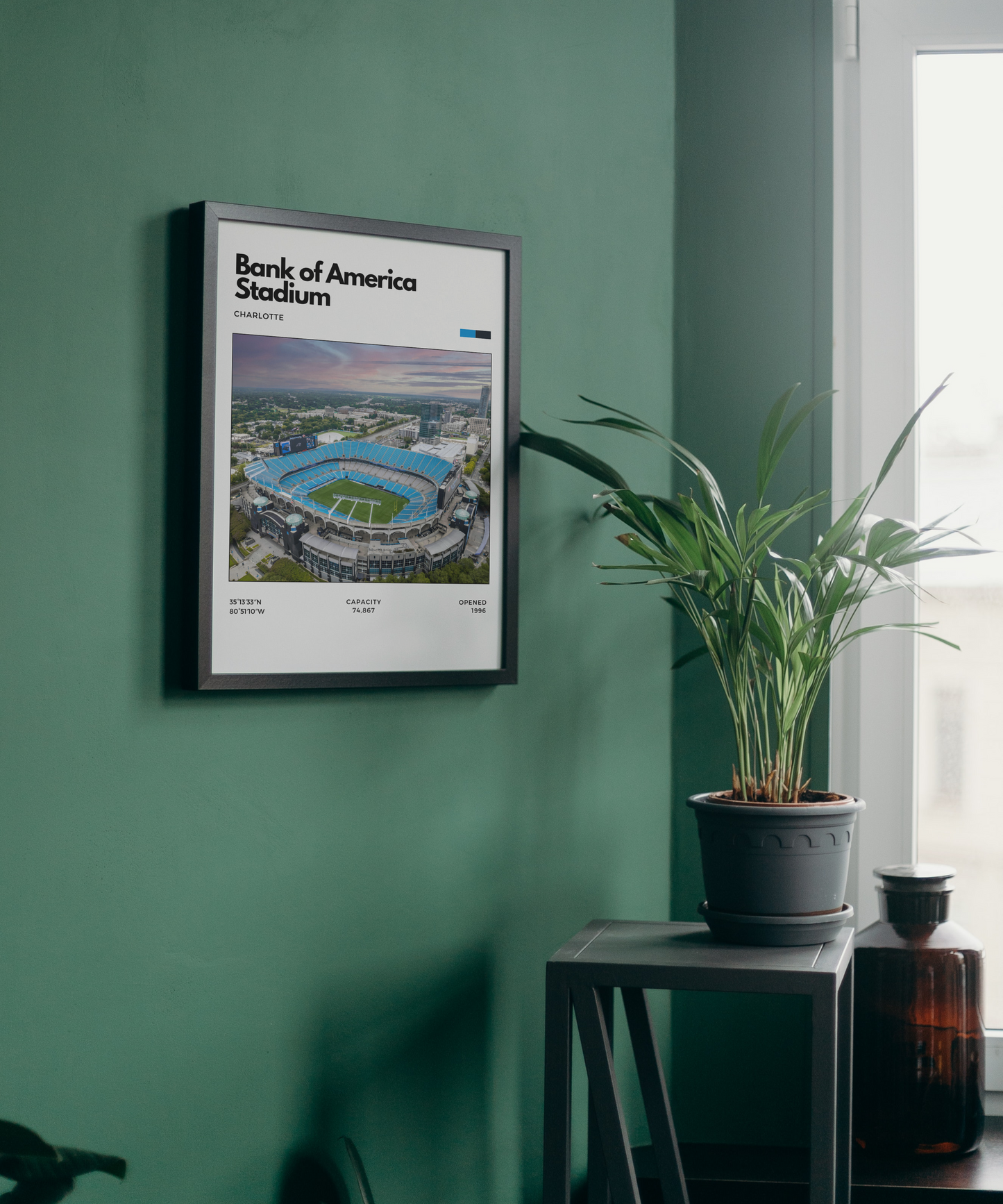 Framed poster of Bank of America Stadium on a green wall with a plant and bottle in the foreground.