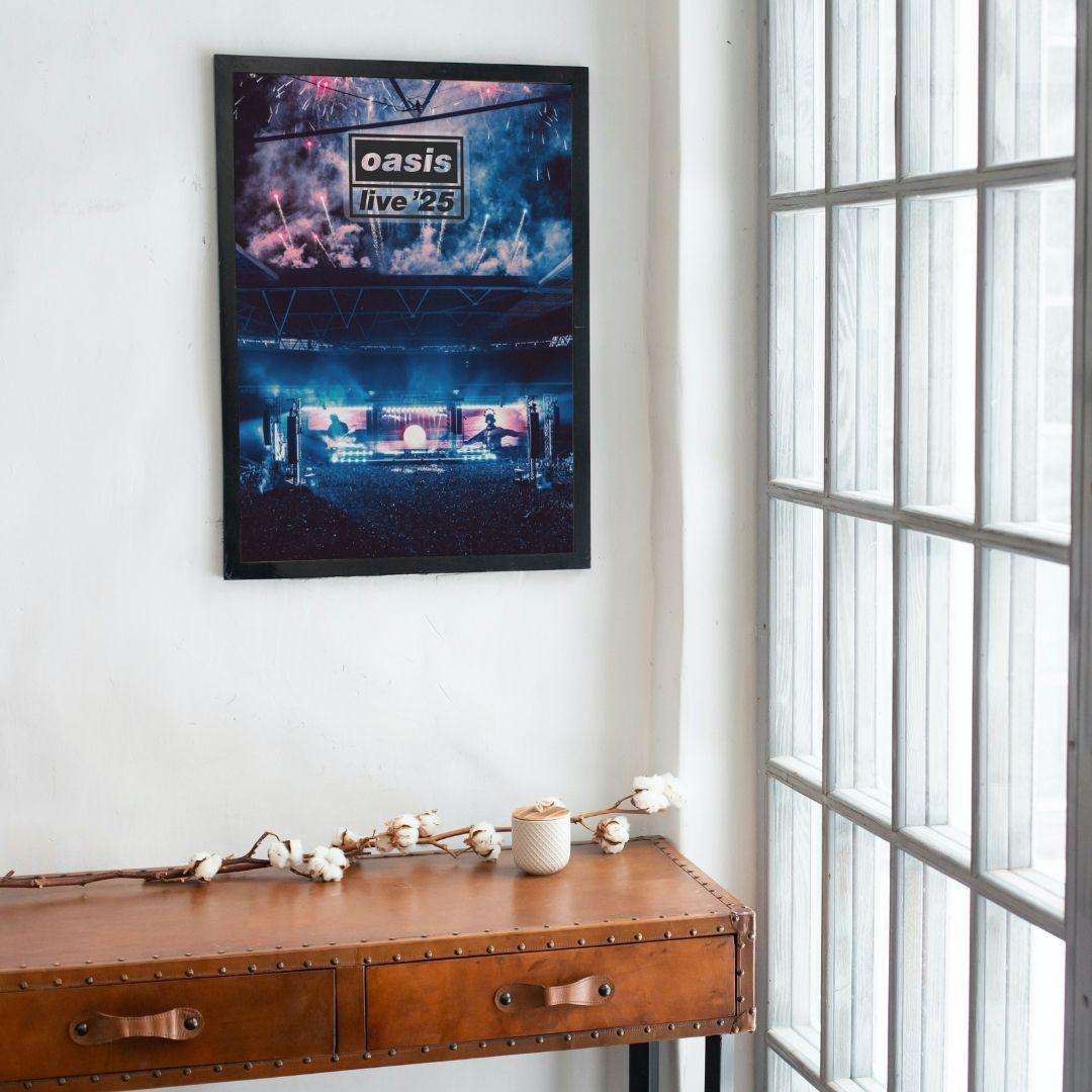 Framed concert poster of Oasis Live 25 Concert Poster. Wembley July 2025 on a wall above a wooden desk with a window in the background.