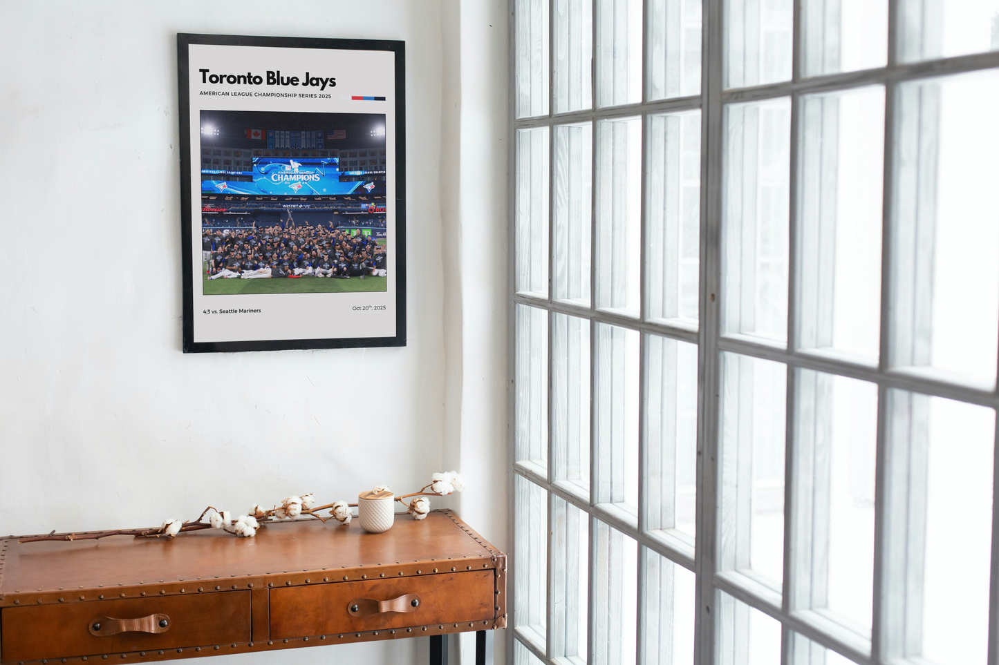 Framed Toronto Blue Jays poster on a wall above a wooden desk with a window in the background.
