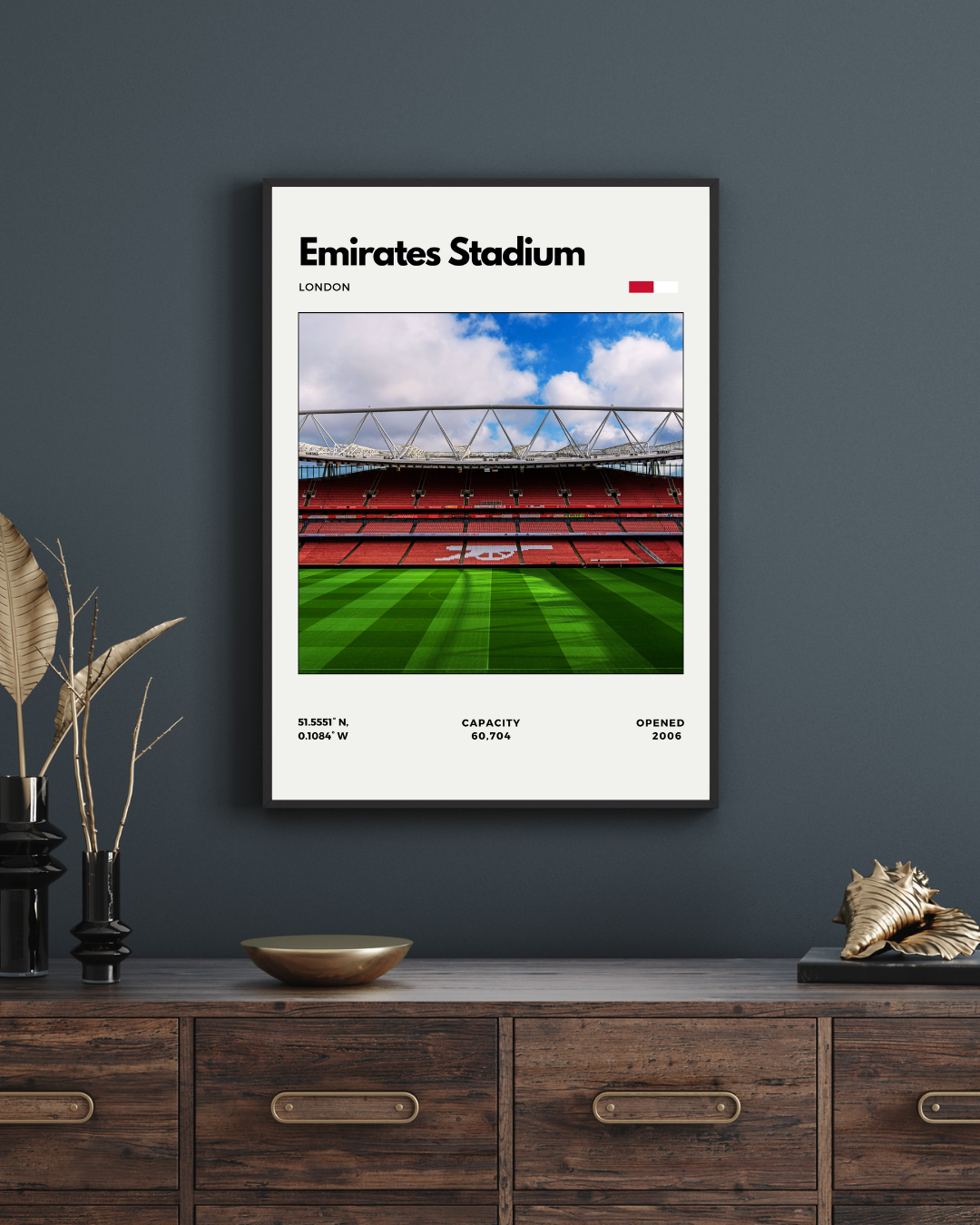 Framed poster of Emirates Stadium on a dark wall above a wooden cabinet.